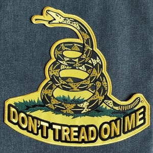 Pode incluir: Patch bordado com uma cascavel enrolada em preto e dourado sobre um fundo amarelo. O patch inclui o texto "DON'T TREAD ON ME" em letras douradas. A cobra está posicionada acima de um tufo de grama verde.