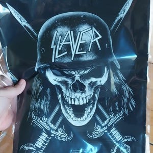May include: Black and white graphic print featuring a skull wearing a helmet with the word "SLAYER" across the front. Two crossed swords are behind the skull. The image is on a black background.