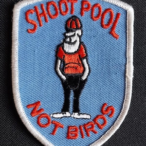 May include: Embroidered patch with a light blue background and a white border. The patch features a cartoon figure in an orange shirt and black pants, with the text "SHOOT POOL NOT BIRDS" in red.