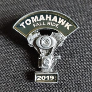 May include: A silver motorcycle engine pin with a black and gold banner that reads "Tomahawk Fall Ride 2019".