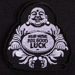 Puede incluir: Un parche bordado en blanco y negro de un Buda sonriente con el texto "Rub Here For Good Luck" en su vientre.