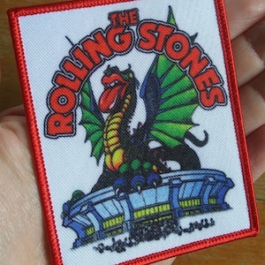 May include: An embroidered patch featuring a cartoon dragon with a tongue sticking out. The dragon is sitting on a blue and yellow structure. The text "The Rolling Stones" is written in a red, white, and black font around the dragon.