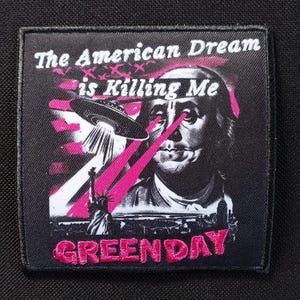 May include: Black square patch with the text "The American Dream is Killing Me" above an illustration of Benjamin Franklin with pink laser beams shooting from his eyes. A UFO and the Statue of Liberty are also featured. The word "GREEN DAY" is embroidered in pink.