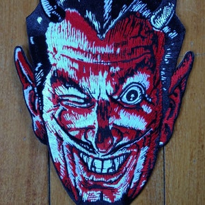 May include: Embroidered patch featuring a devil's face with a winking eye, horns, and a goatee. The design uses red, white, and black thread, creating a detailed and expressive image. The patch is likely for clothing or accessories.