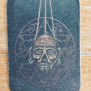 Op de afbeelding: Een zwarte leren patch met een gedetailleerde tekening van een menselijke schedel met een cirkel en een driehoek eromheen. De schedel hangt aan drie touwtjes.