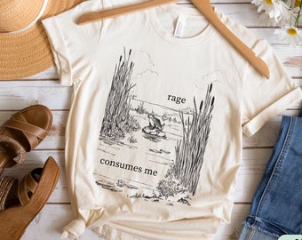 May include: A beige t-shirt with a black and white illustration of a frog in a pond with reeds. The text "rage consumes me" is printed on the shirt.