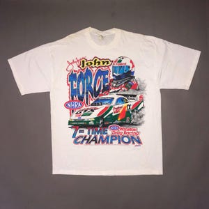 May include: A white short-sleeve t-shirt featuring a graphic print of John Force, a race car, and the text "John Force 7-Time Champion" along with "NHRA Winston Drag Racing". The design includes a driver in a helmet and a green and red race car.