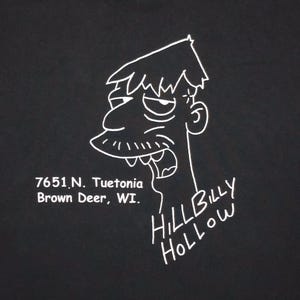 May include: Black t-shirt featuring a white cartoon illustration of a man's face with the text "HILL BILLY HOLLOW" and the address "7651 N. Tuetonia Brown Deer, WI."