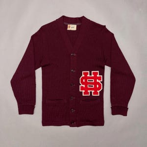 Könnte beinhalten: Eine tiefburgunderrote Strickjacke mit V-Ausschnitt, langen Ärmeln mit gerippten Bündchen und einer Knopfleiste. Auf der unteren linken Seite befindet sich ein großer, kräftig roter Buchstabe S mit weißer Umrandung. Ein kleines weißes Etikett befindet sich am Kragen.