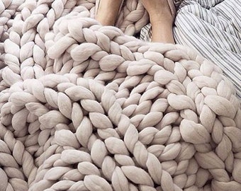 May include: A close-up of a chunky knit throw blanket in a light beige color. The blanket is made of thick, soft yarn and has a relaxed, cozy feel.