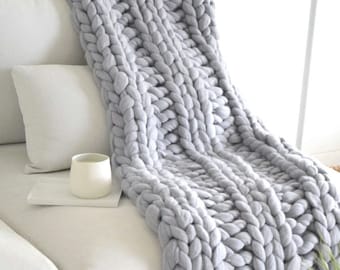 May include: A chunky knit blanket in a light gray color is draped over a white couch. The blanket has a thick, textured weave. A white mug sits on a white tray, next to two white pillows.
