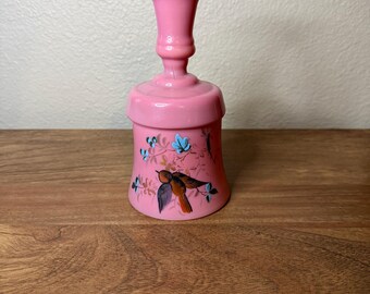 May include: A pink glass bell with a ruffled top and a flared base. The bell features a painted design of a bird and floral elements in blue and black. The bell is displayed on a wooden surface.