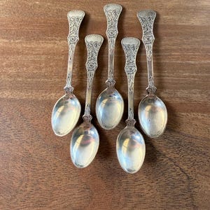 May include: Six antique silver spoons with decorative floral patterns on the handles. The spoons are arranged on a wooden surface, highlighting their polished, reflective bowls. These spoons are likely for serving or dessert.