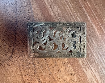 May include: Rectangular silver-toned metal buckle with an ornate, openwork design. The buckle features a scrolling, floral pattern within a rectangular frame. The intricate details and vintage style suggest it may be a belt buckle or decorative element.