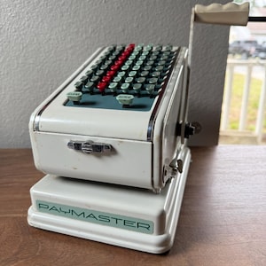 May include: Vintage Paymaster adding machine in off-white with a handle and a series of green and red keys. The word "CLEAR" and "REPEAT" are visible on the machine. The base of the machine has the word "PAYMASTER" printed on it.