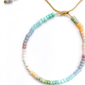 May include: A colorful beaded necklace with a gradient of pastel colors, including light blue, turquoise, green, yellow, pink, and purple. The necklace is tied with a gold cord.