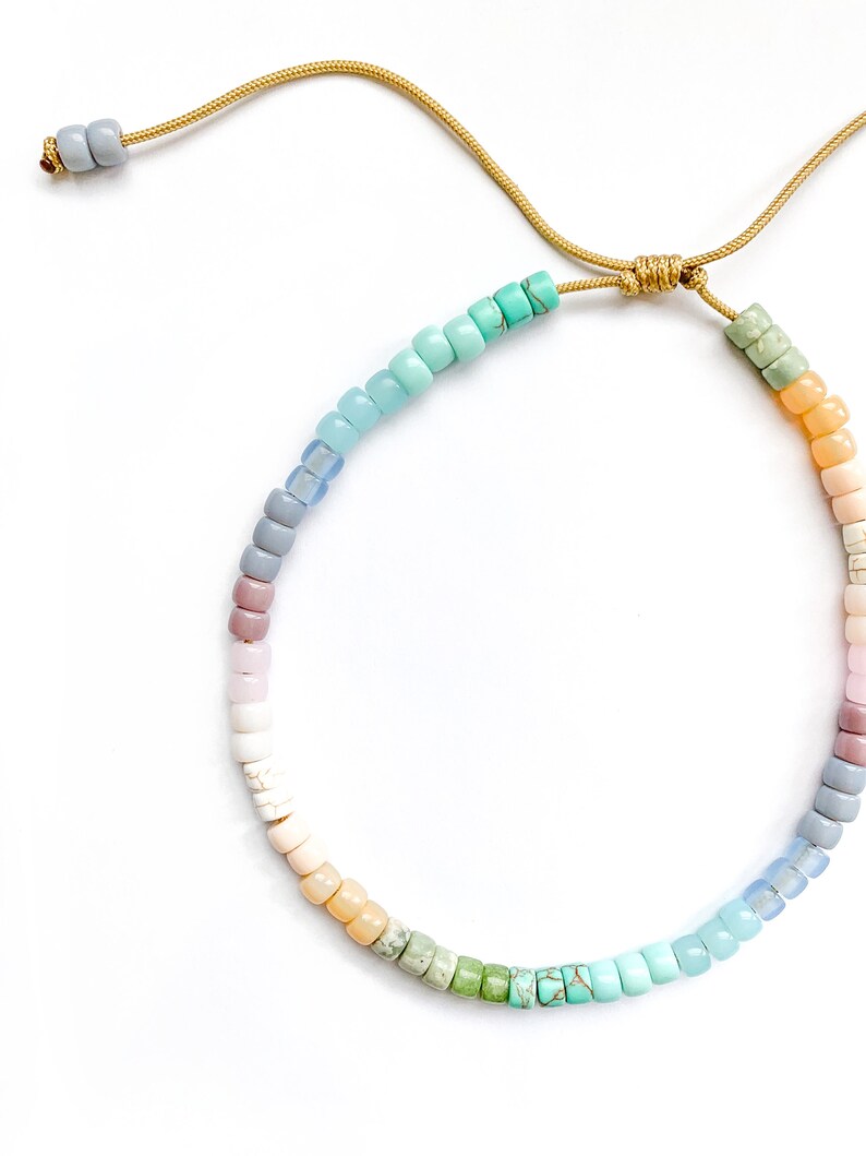 May include: A colorful beaded necklace with a gradient of pastel colors, including light blue, turquoise, green, yellow, pink, and purple. The necklace is tied with a gold cord.