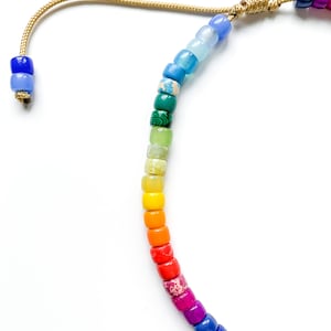 May include: A rainbow colored beaded bracelet with a gold cord tie. The beads are small and round, and they are arranged in a rainbow pattern.