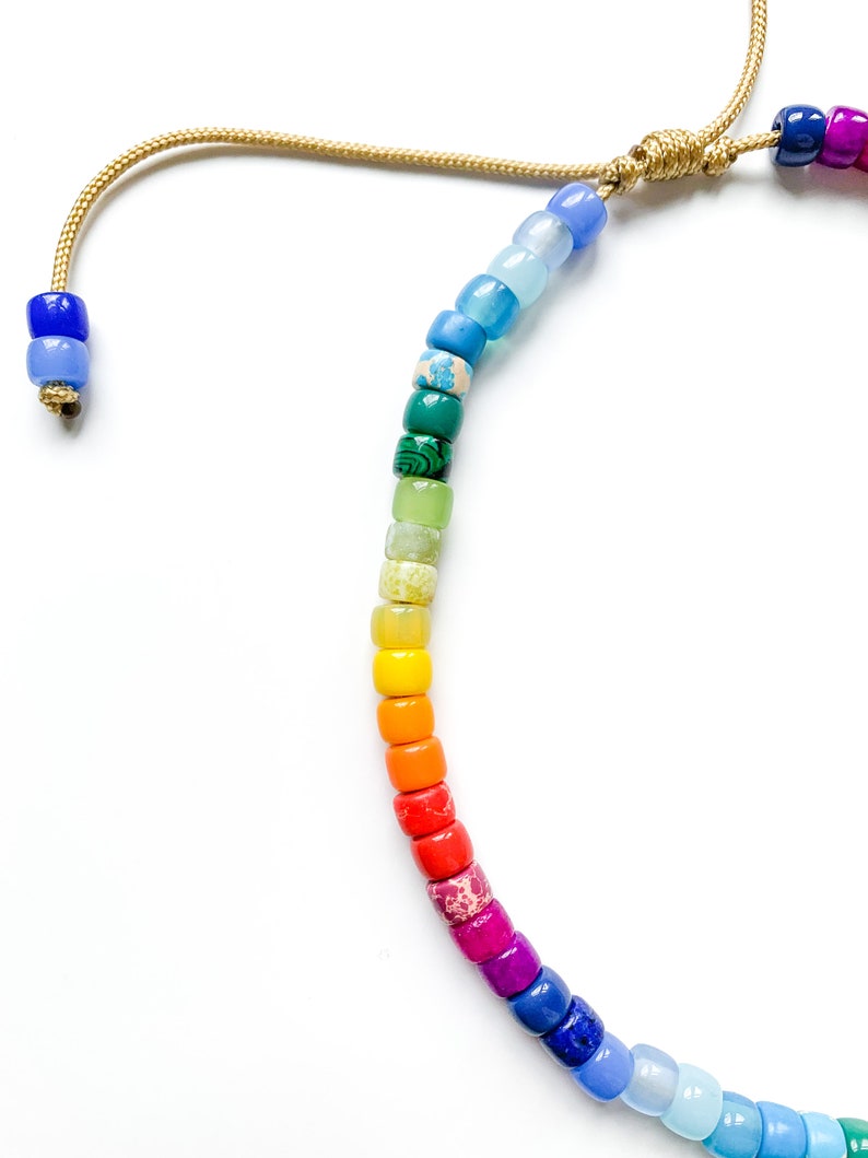 May include: A rainbow colored beaded bracelet with a gold cord tie. The beads are small and round, and they are arranged in a rainbow pattern.