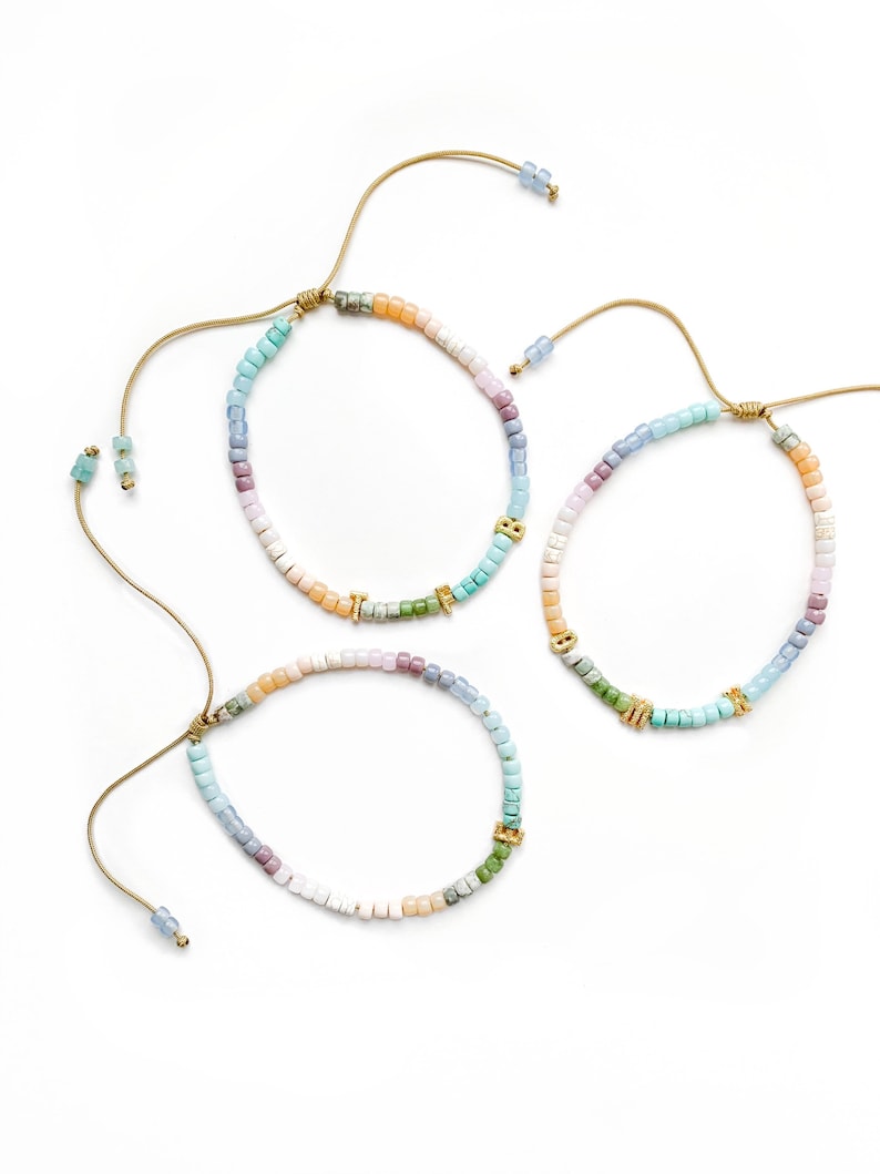 May include: Three colorful beaded bracelets with gold charms. Each bracelet has a different color scheme, with shades of blue, green, pink, and white. The bracelets are tied with a brown cord.