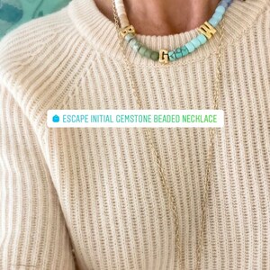 May include: A gold chain necklace with a pendant featuring a pair of stylized wings. The necklace is layered over a multi-colored gemstone beaded necklace with gold letter charms spelling "GWN". The text "ESCAPE INITIAL GEMSTONE BEADED NECKLACE" is visible in the image.