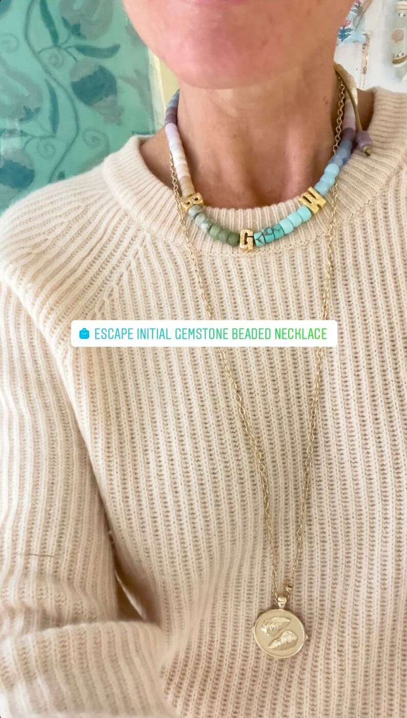 May include: A gold chain necklace with a pendant featuring a pair of stylized wings. The necklace is layered over a multi-colored gemstone beaded necklace with gold letter charms spelling "GWN". The text "ESCAPE INITIAL GEMSTONE BEADED NECKLACE" is visible in the image.
