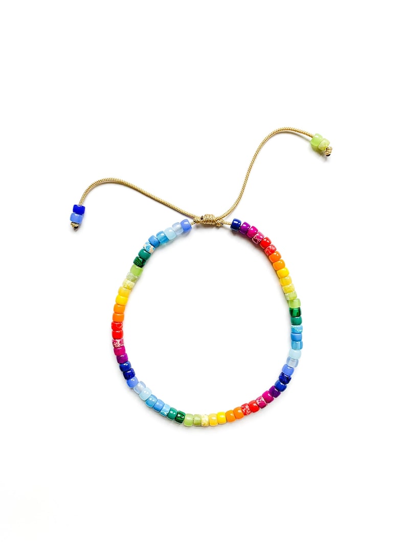 May include: A rainbow colored beaded bracelet with a gold adjustable closure. The bracelet is made of small, square beads in a variety of colors, including red, orange, yellow, green, blue, indigo, and violet.