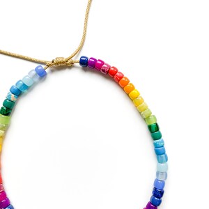 May include: A rainbow colored beaded bracelet with a gold cord closure. The beads are small and round, and they are arranged in a rainbow pattern.