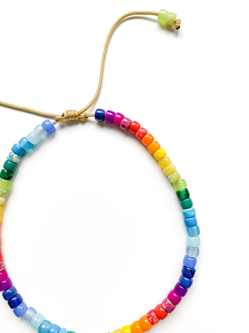 May include: A rainbow colored beaded bracelet with a gold cord closure. The beads are small and round, and they are arranged in a rainbow pattern.
