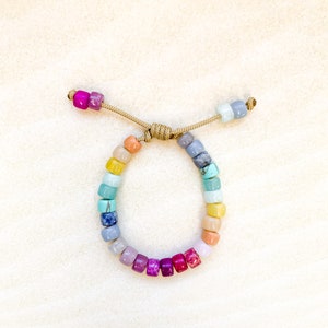 May include: A colorful beaded bracelet with a rainbow of colors, including yellow, orange, red, purple, blue, and green. The bracelet is made with small, square-shaped beads and is tied with a brown cord.