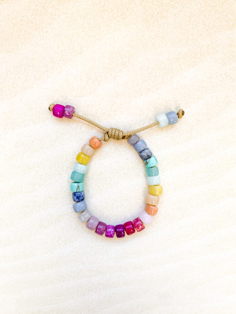 May include: A colorful beaded bracelet with a rainbow of colors, including yellow, orange, red, purple, blue, and green. The bracelet is made with small, square-shaped beads and is tied with a brown cord.
