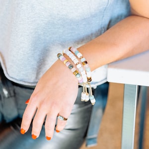 May include: A woman's wrist adorned with four layered bracelets. The bracelets are made of white, pink, blue, and brown beads with gold accents. The bracelets are tied with a gold cord.
