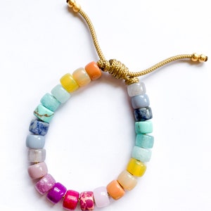 May include: A rainbow colored beaded bracelet with a gold cord closure. The beads are made of various semi-precious stones and are arranged in a gradient pattern.