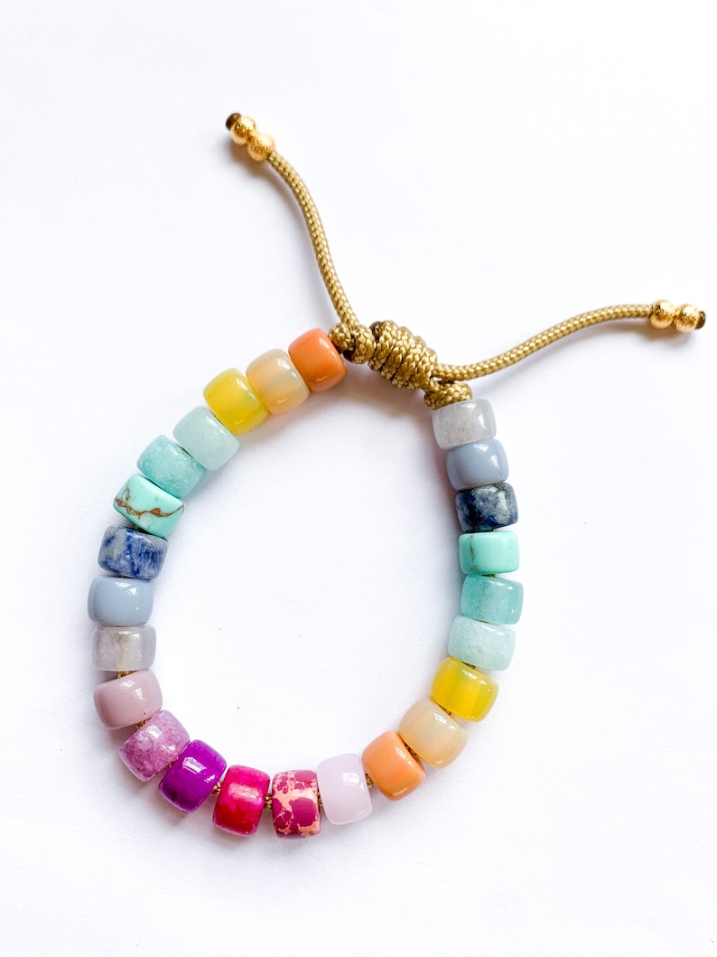May include: A rainbow colored beaded bracelet with a gold cord closure. The beads are made of various semi-precious stones and are arranged in a gradient pattern.