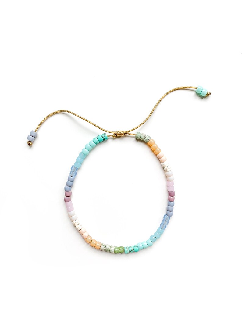 May include: A colorful beaded bracelet with a rainbow gradient of pastel blue, green, pink, and orange beads. The bracelet is tied with a thin gold cord.