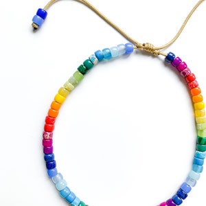May include: A rainbow colored beaded bracelet with a gold cord closure. The beads are small and round, and they are arranged in a rainbow pattern.