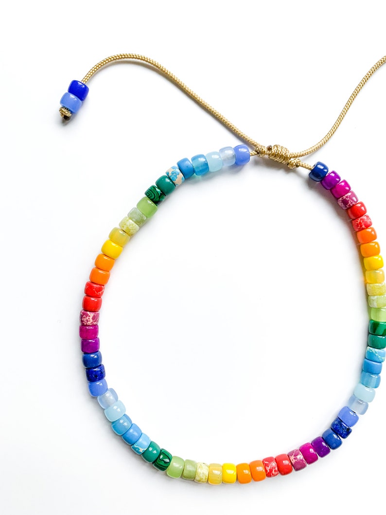 May include: A rainbow colored beaded bracelet with a gold cord closure. The beads are small and round, and they are arranged in a rainbow pattern.