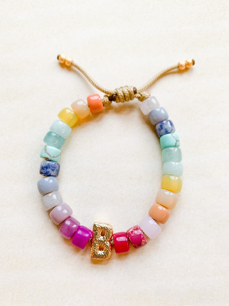 May include: A rainbow beaded bracelet with a gold letter "B" charm. The beads are in shades of blue, green, yellow, orange, pink, and purple.