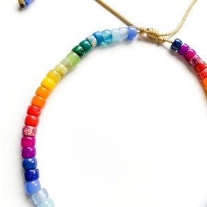 May include: A rainbow-colored beaded necklace with a gold cord. The beads are small and round, and they are arranged in a rainbow pattern. The necklace is tied with a knot at the top.