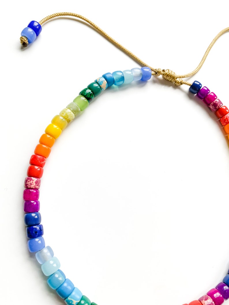 May include: A rainbow-colored beaded necklace with a gold cord. The beads are small and round, and they are arranged in a rainbow pattern. The necklace is tied with a knot at the top.