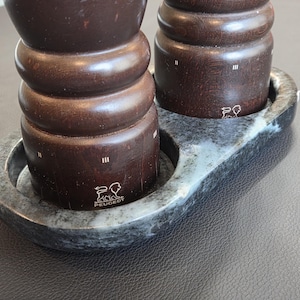 May include: Two dark brown wooden pepper and salt mills with horizontal ridges, featuring the Peugeot lion logo and Roman numerals II and III, resting in a gray and white mottled stone tray with two circular indentations for the mills.