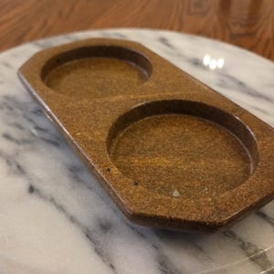 May include: A brown, elongated wooden tray featuring two circular, recessed compartments. The tray has a smooth, polished finish with a visible wood grain texture and beveled edges, designed to hold two round items securely in place.
