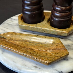 May include: A polished stone tray with a tan and brown speckled pattern, a matching stone holder containing two dark brown wooden pepper mills, and a large circular white marble board with grey veining underneath the items.