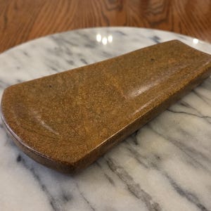 May include: A rectangular, slightly curved brown tray with a smooth, speckled finish. The object features a shallow, elongated basin with rounded edges and a polished surface, showcasing a consistent earthy tone throughout its entire structure.