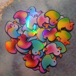 May include: A collection of iridescent rainbow rubber duckies in various poses. The ducks are made of a shiny, reflective material and have a holographic finish.