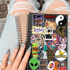May include: A black notebook with a variety of colorful stickers, including a yin yang symbol, a pizza slice, a cartoon character, a game cartridge, and a lava lamp. The notebook has a spiral binding and is covered in stickers.