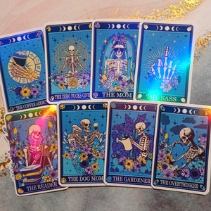 May include: Set of ten holographic tarot cards with colorful floral designs and skeletons. The cards feature titles such as "The Coffee Addic", "The Zero Fucks Give", "The Mom", "The Sass", "The Dog Mom", "The Gardener", "The Overthinker", and "The Reader".