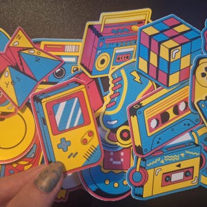 May include: A collection of colorful stickers featuring retro designs, including a cassette tape, a game console, a floppy disk, a roller skate, a smiley face, and a Rubik's cube.