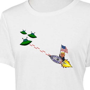 Könnte beinhalten: Weißes T-Shirt mit einem Cartoon-Bild einer Person, die eine amerikanische Flagge hält und auf einer Rakete reitet, die einen Laserstrahl auf drei fliegende Untertassen schießt. Der Text auf der Rakete lautet "Space Force".