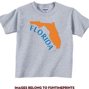 Könnte beinhalten: Hellgraues T-Shirt mit orangefarbener Umrisszeichnung des Bundesstaates Florida und dem Wort "FLORIDA" in Blau. Der Text ist gebogen, um sich der Form des Staates anzupassen. Das Shirt ist aus weichem Material und für Kinder geeignet.